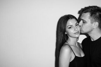 Чёрно-белая фотосессия пары в стиле нулевых: парень целует девушку в щёку у светлой стены, вспышка