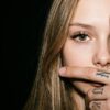 Фото глаза крупным планом: лицо девушки закрывает мужская рука с татуировками на пальцах, чёрный фон